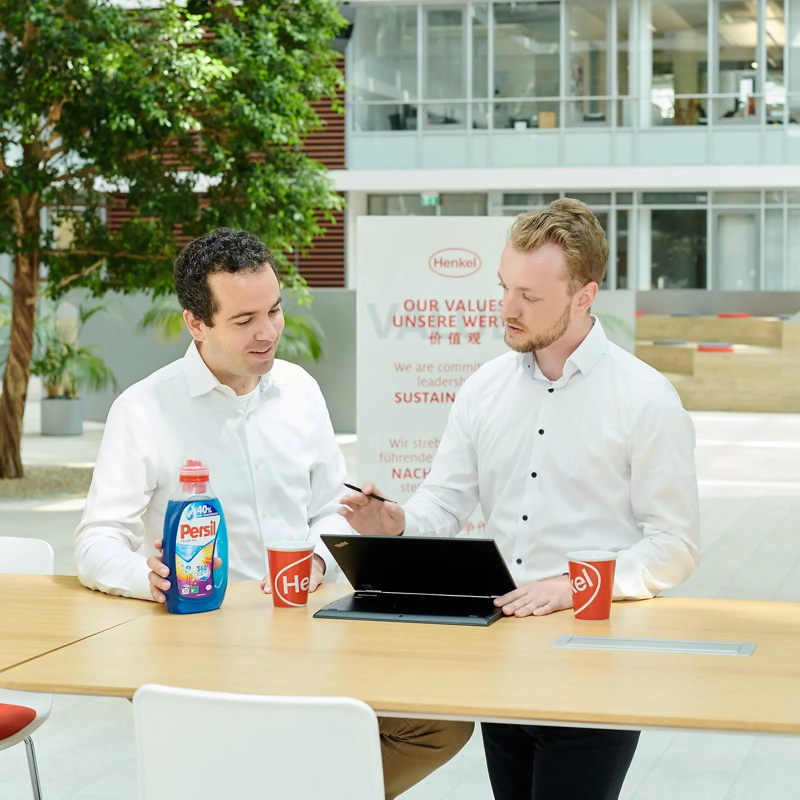 Két férfi egy asztal mellett áll egy Persil flakonnal és egy laptoppal