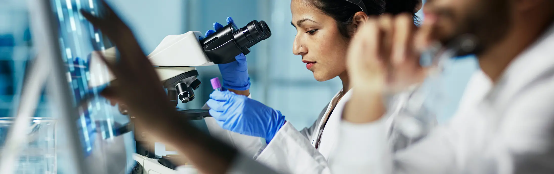 egy laboratóriumban egy nő ül egy mikroszkóp előtt, amint egy mintát vizsgál, elöl pedig egy szakállas férfi nézi a képernyőt.