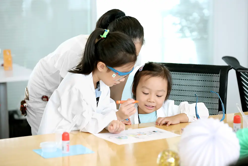 Két kisgyerek egy kutatónővel képet színeznek az asztalnál