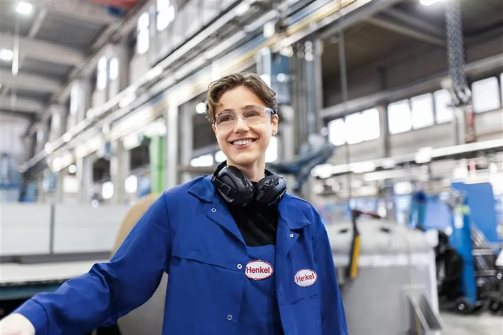 Egy mosolygó Henkel-munkatárs kék munkakabátban és biztonsági fejhallgatóban áll egy ipari gyártócsarnokban, a gyártás szakértelmét és innovációját képviselve.