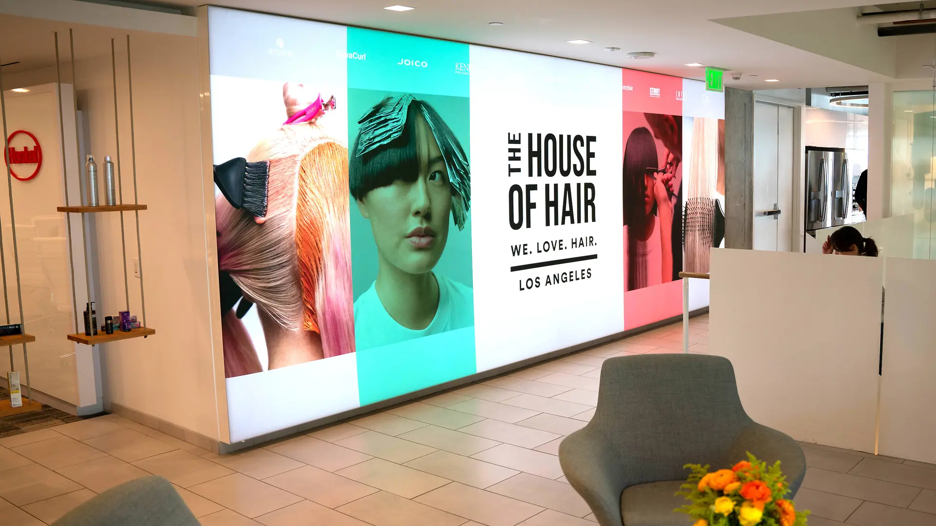 Modern előcsarnok fényes fali kijelzővel, amelyen nagy, színes, haj tematikájú kép és a „THE HOUSE OF HAIR – WE. LOVE. HAIR. – LOS ANGELES.” felirat láthatók. A minimalista kialakítású, járólapos, fehér térelválasztókkal tagolt belső térben világosszürke lounge-székek és kis asztal élénk virágkompozícióval.