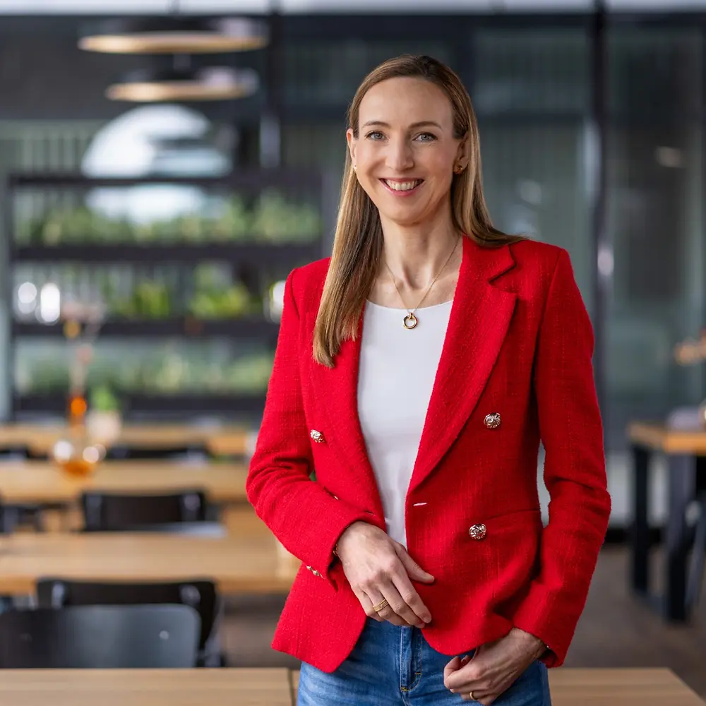 A fehér felsőt és élénk piros blézert viselő Dr. Simone Bagel-Trah modern kávéházi hangulatú környezetben, háttérben fa asztalokkal és vázákkal.