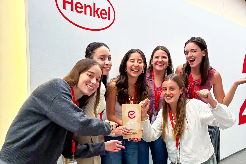 Hat emberből álló csoport áll együtt a Henkel logóval ellátott fal előtt. Piros nyakpántokat és hétköznapi ruhákat viselnek, és csoportba gyűlnek egy díjplakett körül. Úgy tűnik, hogy a helyszín egy beltéri vállalati tér világos színű fallal és szürke padlóval. A csoport lelkesen pózol, kiemelik a középen látható díjat.