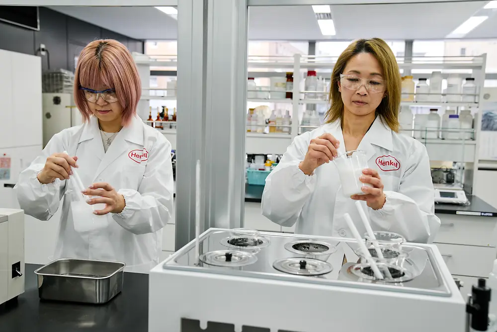 Két fehér Henkel-laboratóriumi köpenyt viselő személy dolgozik egy modern laboratóriumban. Tudományos műszereket és edényeket kezelnek egy pulton, a háttérben pedig különböző laboratóriumi berendezések és a felszereléseket tartalmazó polcok láthatók.