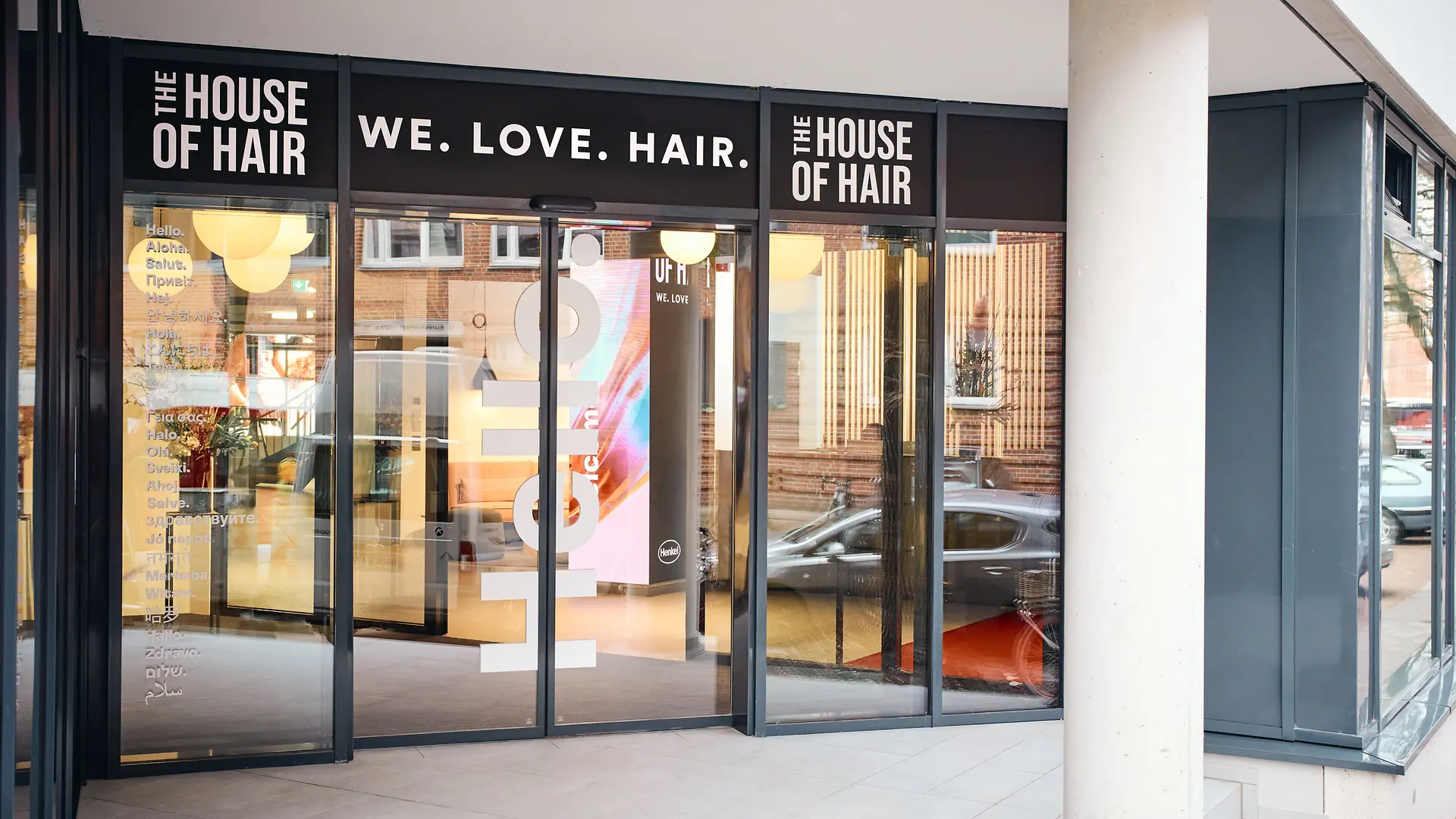 Üvegezett bejárat, az ajtó felett a „We. Love. Hair.” szlogen olvasható. Jobbra és balra tőle a „The House of Hair” felirat látszik.