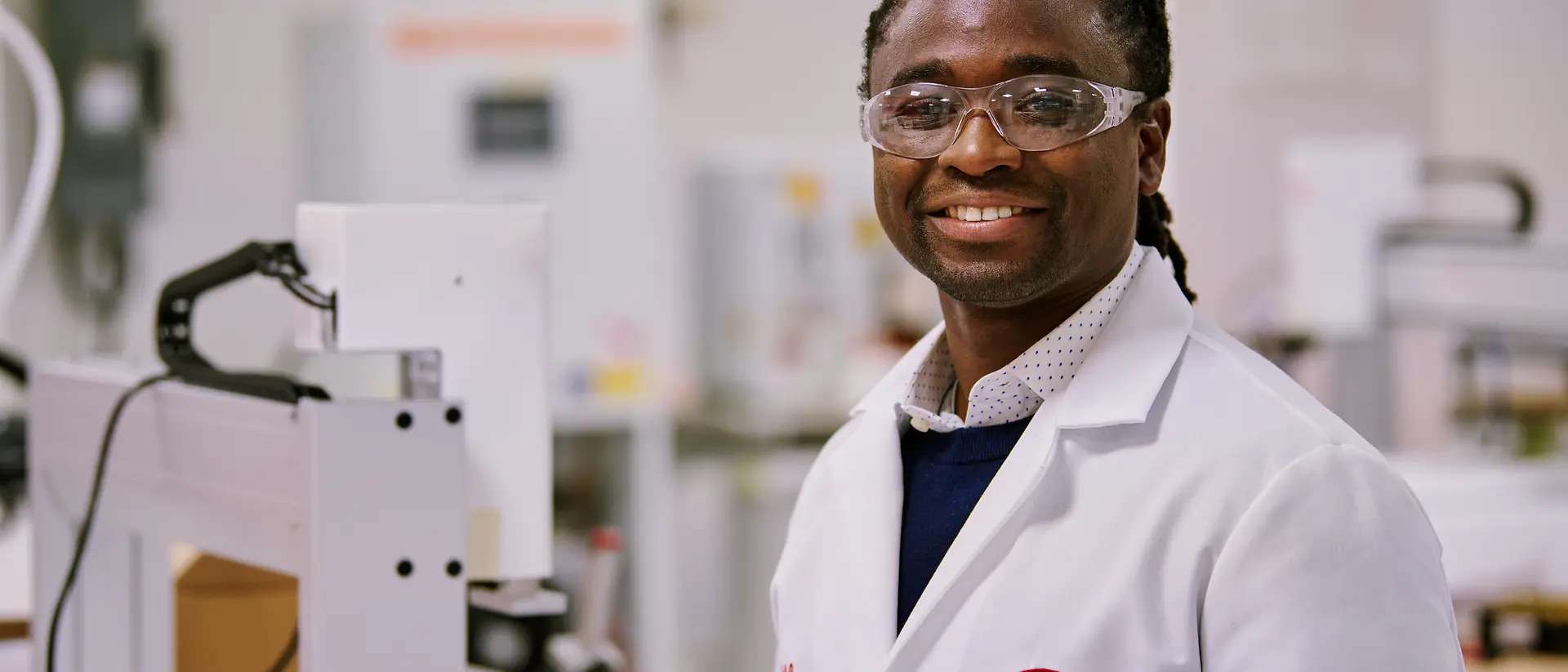 Henkel feliratú, fehér laboratóriumi köpenyt viselő személy laborkörnyezetben, berendezések és gépek között.