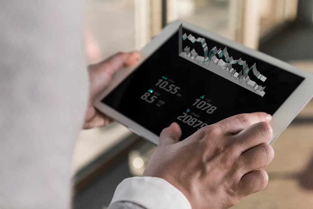 Váll feletti nézetből, világos beltéri környezetben ábrázolt személy kezében digitális grafikonokat és számadatokat megjelenítő táblagéppel.